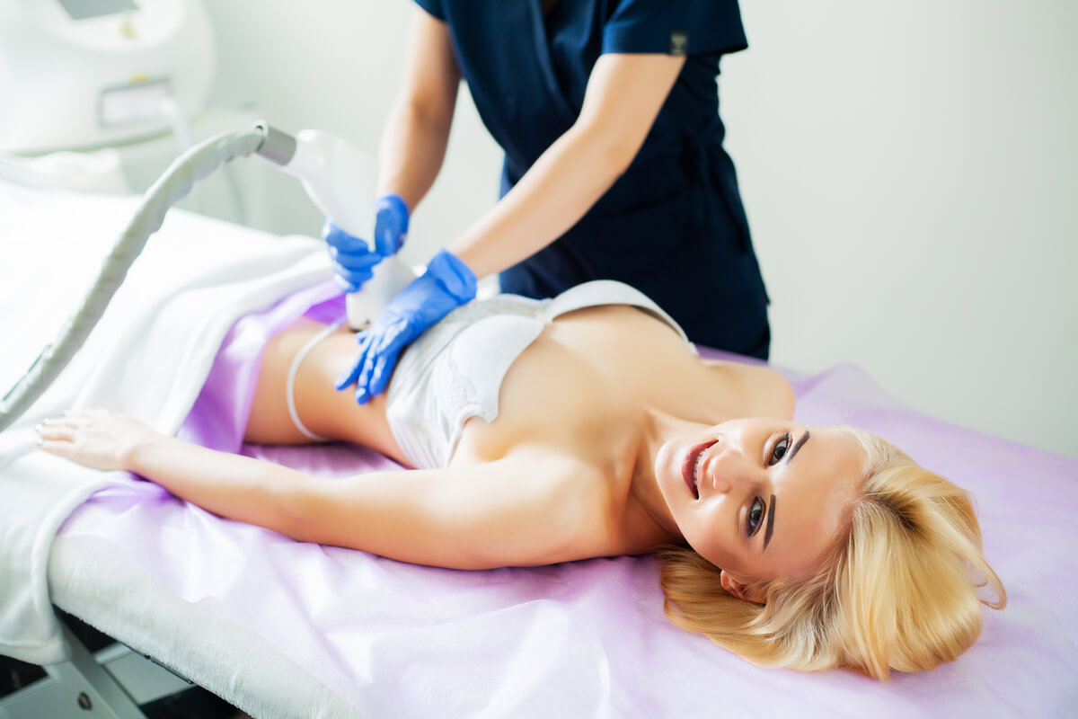 A patient is receiving a non-invasive body contouring treatment at a medical spa. The procedure involves a practitioner using a specialized device to target areas of the patient's midsection. The patient, who is lying comfortably on a treatment bed and dressed in a white bandeau and underwear, smiles contentedly. The practitioner wears gloves and is focused on the procedure, applying the device to the patient's abdomen. The setting is a clean, clinical environment, indicating a professional and controlled atmosphere for the body contouring process.