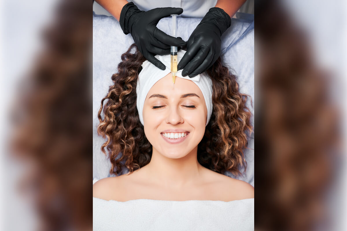 Botox - A woman is lying down with her eyes closed, smiling as a cosmetic professional prepares to administer a Botox injection into her forehead. The professional is wearing black gloves and holding a syringe, carefully targeting the area between the woman’s eyebrows. The woman is wearing a white headband to keep her hair back, and she is covered with a white towel, indicating a clinical or spa-like setting. This Botox procedure aims to reduce fine lines and wrinkles, providing a smoother and more youthful appearance. The overall atmosphere suggests relaxation and professional care during this cosmetic treatment.