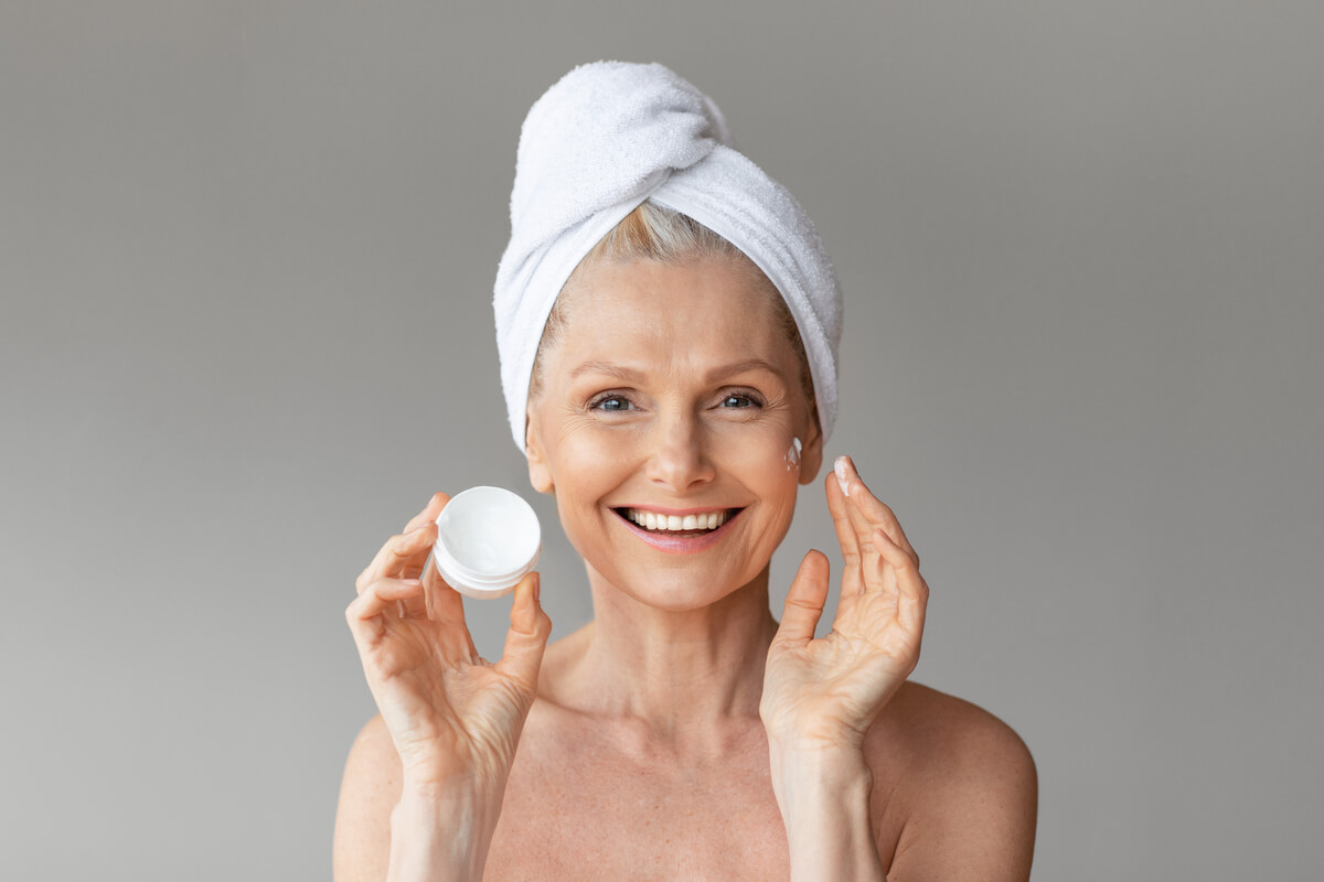 Anti-Ageing Skincare - An older woman with a towel wrapped around her head is smiling as she applies an anti-ageing cream to her face. She holds a small jar of the cream in one hand, while the other hand gently touches her cheek, where a small dab of cream is visible. Her radiant smile and clear complexion highlight the positive effects of her skincare routine. The neutral background emphasizes the simplicity and focus on self-care. This image conveys a message of joy and confidence in maintaining youthful, healthy skin through the use of effective anti-ageing skincare products.