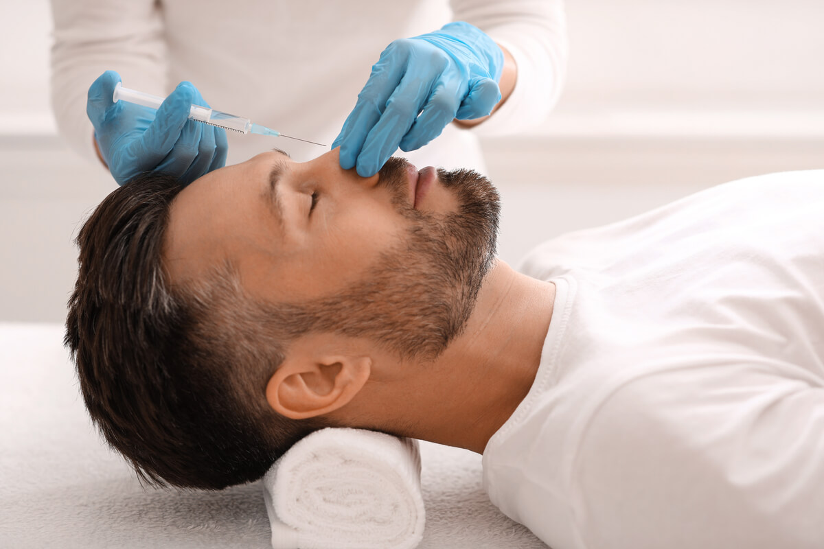 Non-Surgical Rhinoplasty - A man with a neatly trimmed beard is lying on a treatment bed, receiving a non-surgical rhinoplasty procedure in a professional aesthetic clinic. The practitioner, wearing blue gloves, is precisely injecting dermal fillers into the bridge of the man's nose using a syringe. The man is resting his head on a rolled towel, with his eyes closed, appearing calm and relaxed. The clinic's bright, clean environment emphasizes the safe and non-invasive nature of the procedure, which aims to refine the nose shape and enhance facial symmetry without surgery.
