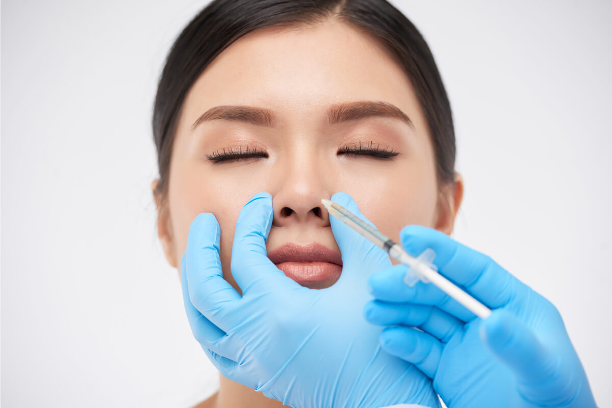 Non-Surgical Rhinoplasty - A young woman is undergoing a non-surgical rhinoplasty procedure in a clinical setting. The image shows her with her eyes closed as a practitioner, wearing blue gloves, carefully injects dermal fillers into the tip of her nose using a syringe. The treatment aims to refine the shape of her nose, correcting minor imperfections and enhancing her facial symmetry without the need for invasive surgery. The setting is sterile and professional, highlighting the focus on precision and safety in this aesthetic enhancement procedure.